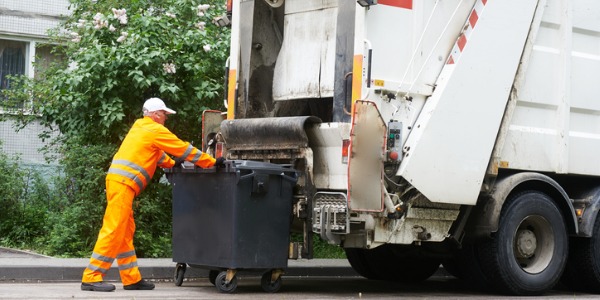 A garbage collector loading a garbage bin onto the lift of a garbage truck.