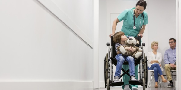 An orderly pushing a child in a wheelchair through a hospital corridor.