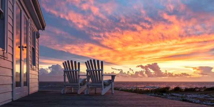 Two adirondack chairs on a vacation home deck, facing a beautiful sunset.