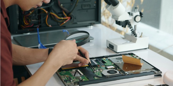 A desktop computer technician examining a motherboard with a magnifying glass while sitting at his desk.