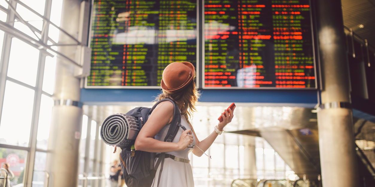 A young woman in a dress and backpack looking at flight information in the airport terminal.