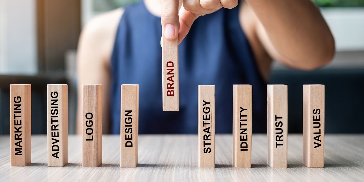 A brand strategy concept - a hand placing wooden dominos on a table. Each domino has text - Marketing, Advertising, Logo, Design, Brand, Strategy, Identity, Trust and Values.