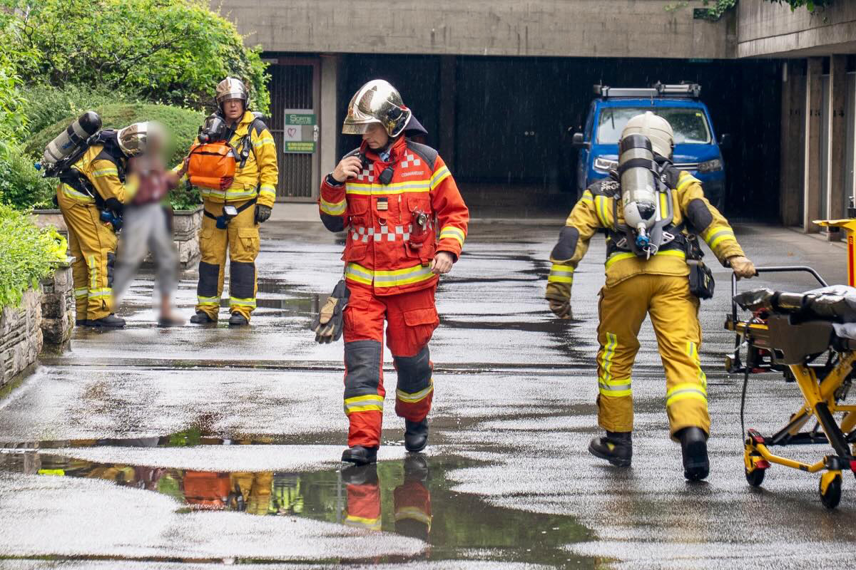 Mehrere Feuerwehrleute in gelben und roten Anzügen sind vor einem Gebäude im Einsatz. Eine Person in Zivil ist ebenfalls zu sehen.