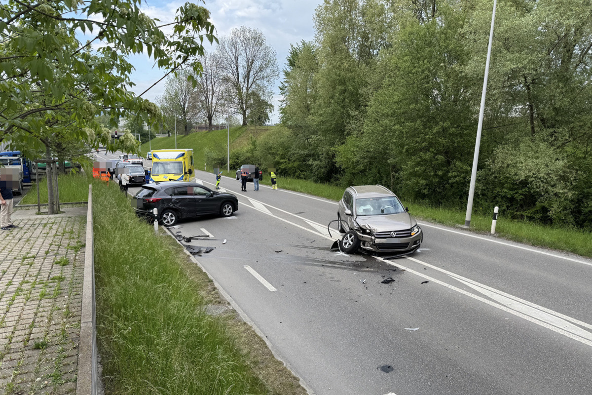Zu sehen sind zwei beschädigte Autos nach einem Unfall auf einer zweispurigen Straße. Ein Rettungsfahrzeug ist im Hintergrund zu erkennen.