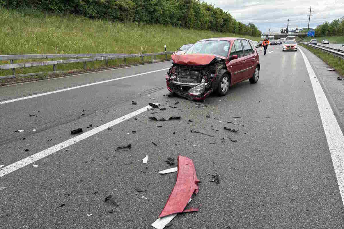 Auf der Fahrbahn steht ein rotes Auto mit stark beschädigter Front. Um das Auto liegen verstreute Trümmerteile, während ein weiteres Auto auf die Szene zukommt.