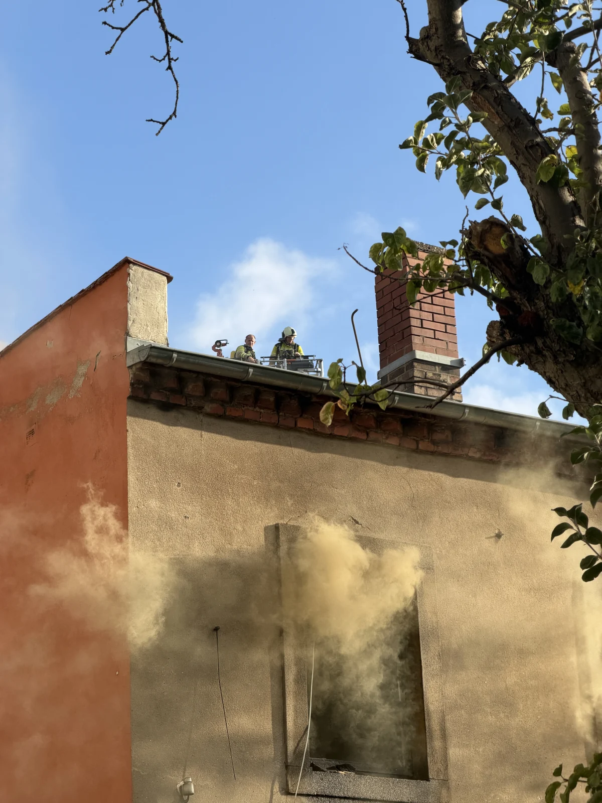 Rauch dringt aus dem Fenster eines mehrstöckigen Gebäudes, während Feuerwehrleute auf dem Dach Arbeiten ausführen. Ein Baum ist links zu sehen.