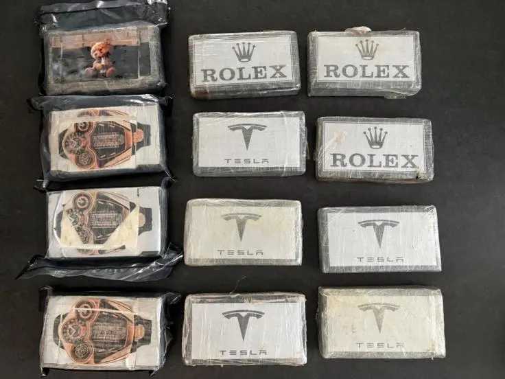 Auf einer dunklen Oberfläche liegen elf in Plastik verpackte Pakete, die Logos von Rolex und Tesla tragen.