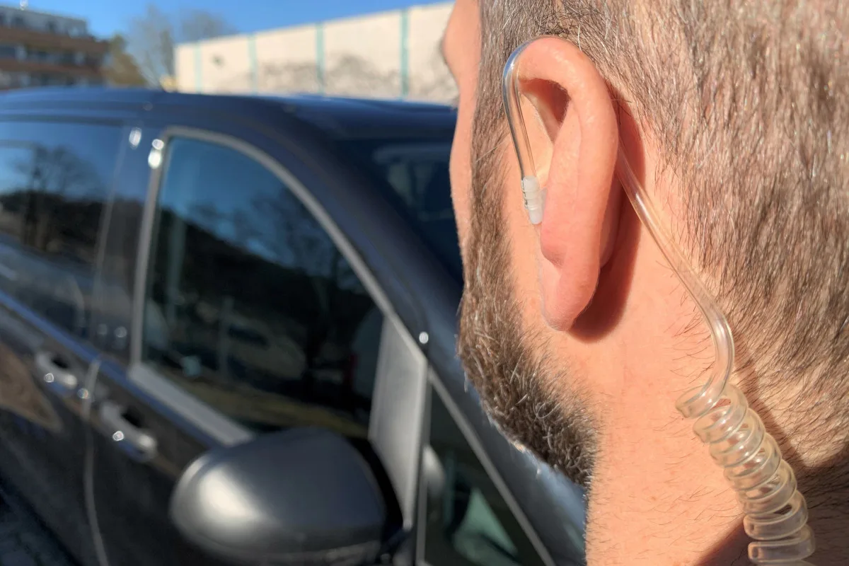 Ein Mann trägt einen transparenten Kommunikations-Ohrhörer. Im Hintergrund ist ein schwarzes Auto sichtbar.