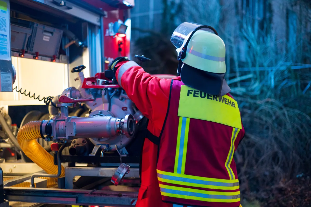 Ein Feuerwehrmann in Schutzkleidung und Helm bedient eine Wasserpumpe an einem roten Einsatzfahrzeug. Die Umgebung ist dunkel, beleuchtet von den Fahrzeuglichtern.