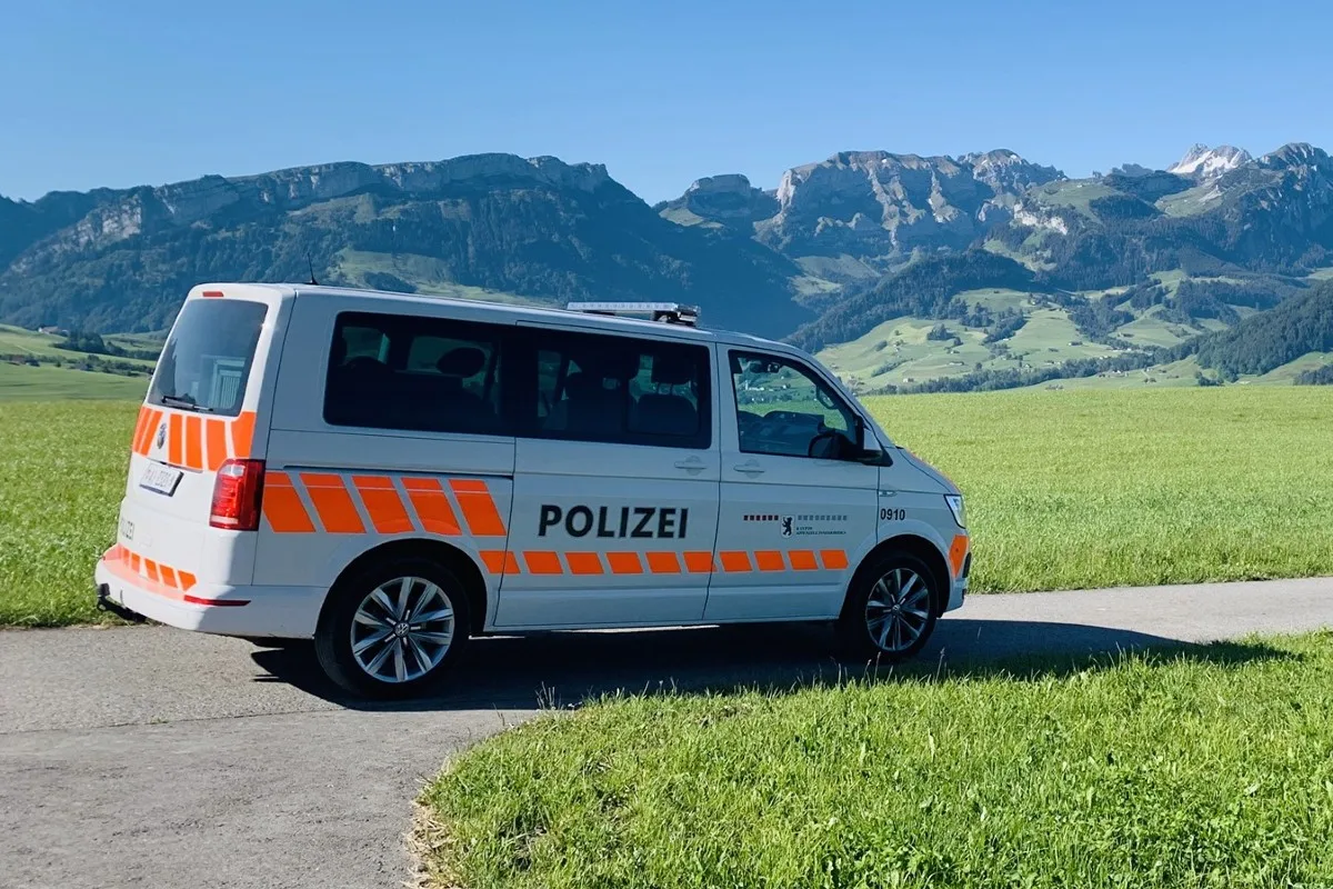 Ein weißer Polizeivan mit orangenen Streifen und der Aufschrift 'Polizei' steht auf einer Landstraße. Im Hintergrund sind grüne Wiesen und Berge zu sehen.