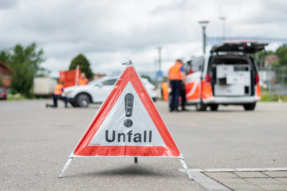 Ein Dreiecksschild mit 'Unfall' steht im Vordergrund, während im Hintergrund Rettungskräfte arbeiten.