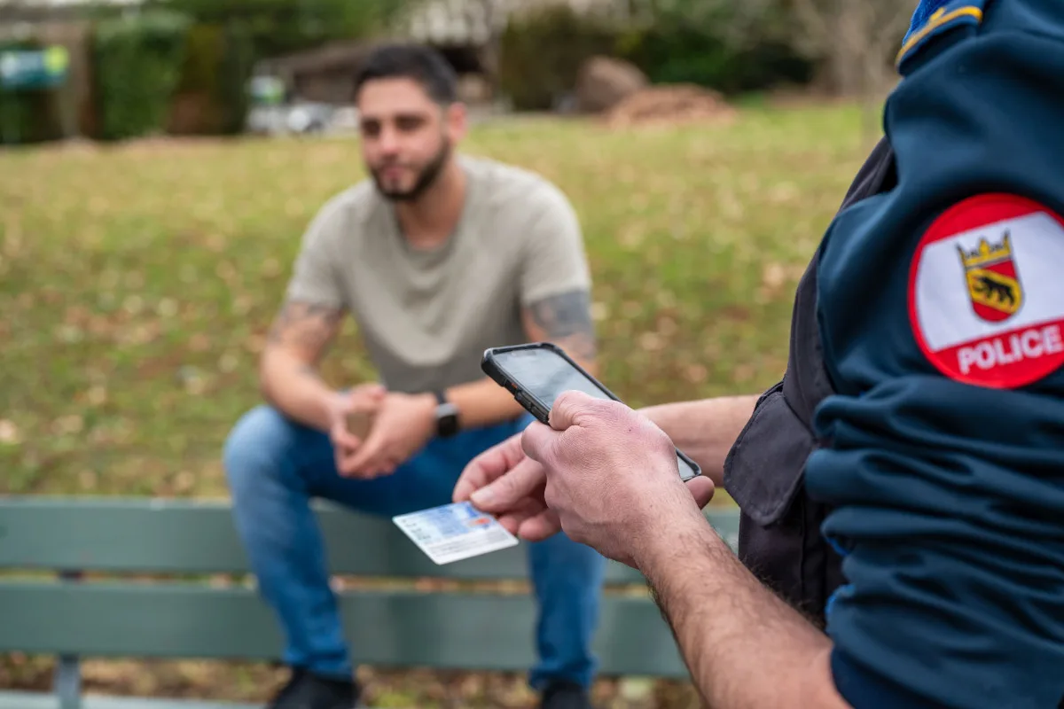 Ein Polizist mit Uniform überprüft einen Personalausweis, während ein Mann auf einer Parkbank sitzt. Das Bild zeigt eine Parksituation mit zivilem Hintergrund.