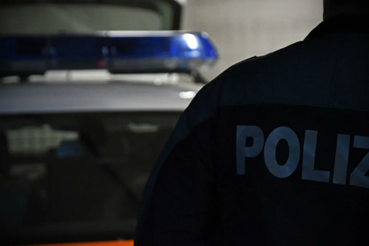 Ein Polizist steht in Uniform, während im Hintergrund ein Polizeifahrzeug mit Blaulicht erscheint. Es ist dunkel oder dämmernd.