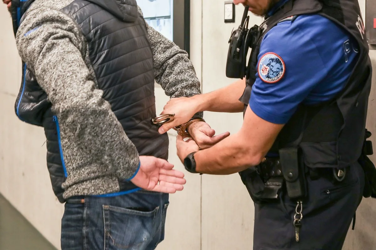 Ein Polizist in blauer Uniform legt einer Person in grauer Jacke Handschellen an. Sie befinden sich in einem Innenraum.