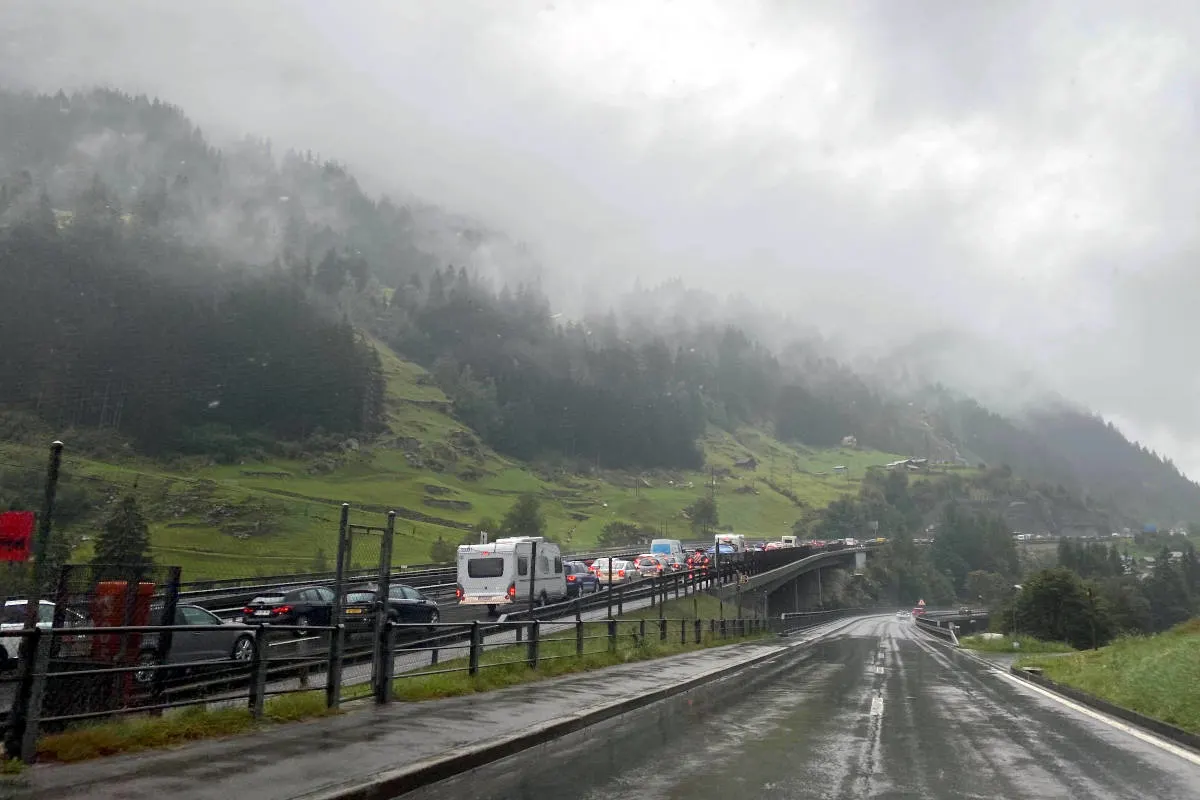 Eine lange Reihe von Fahrzeugen steht im Stau auf einer nassen Autobahnbrücke. Im Hintergrund ist eine neblige Berglandschaft zu sehen.