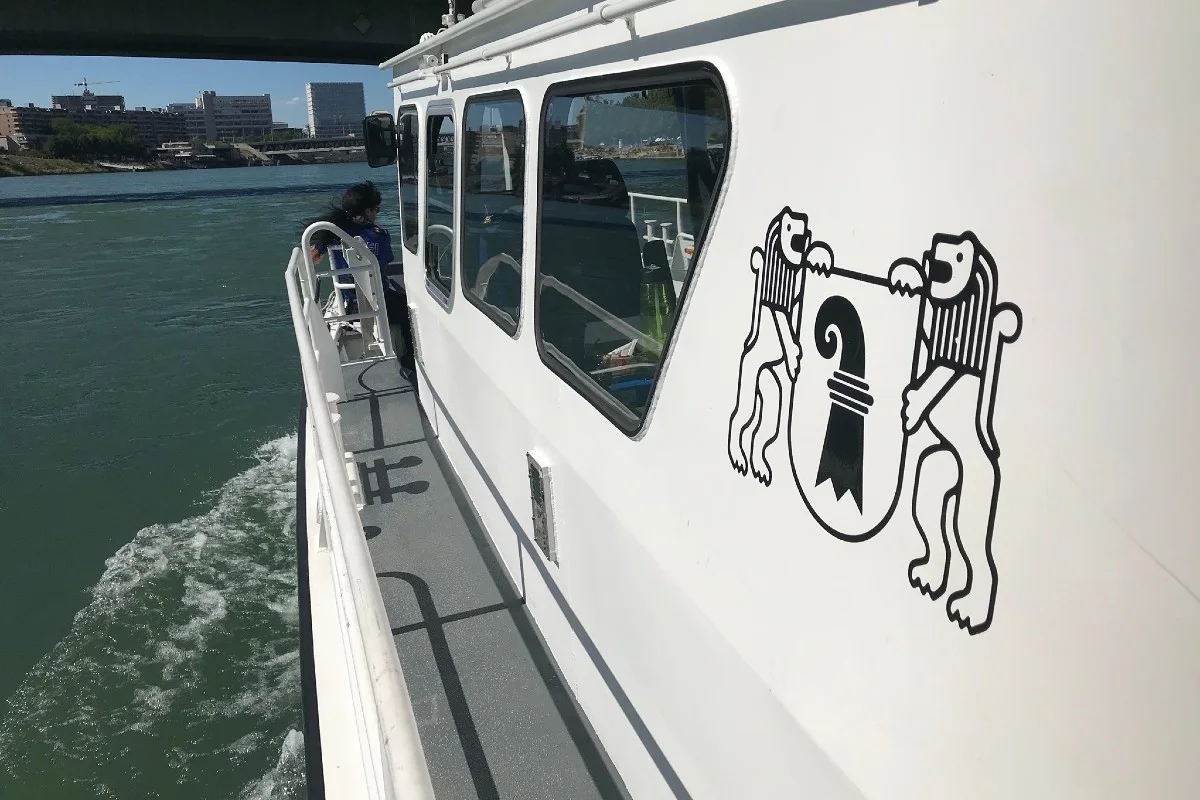 Ein Polizeiboot mit dem Wappen des Kantons Basel-Stadt fährt auf einem Fluss unter einer Brücke.