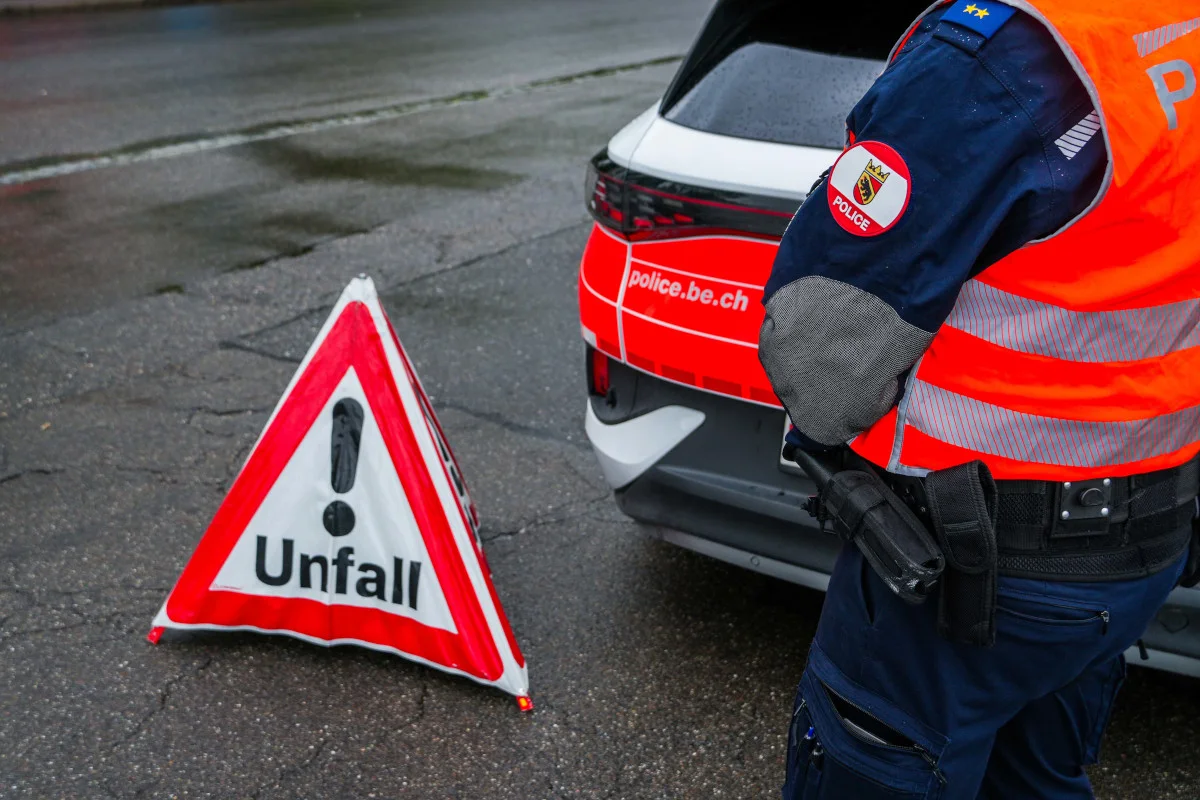 Ein Polizist in Uniform steht neben einem Unfall-Warnschild auf einer nassen Straße. Im Hintergrund ist ein Polizeifahrzeug der Berner Polizei zu sehen.