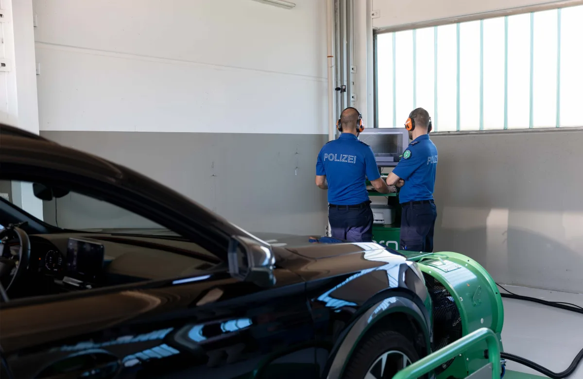 Zwei Polizisten in blauer Uniform stehen in einer Garage vor technischen Geräten. Ein schwarzes Auto ist nahe sichtbar.