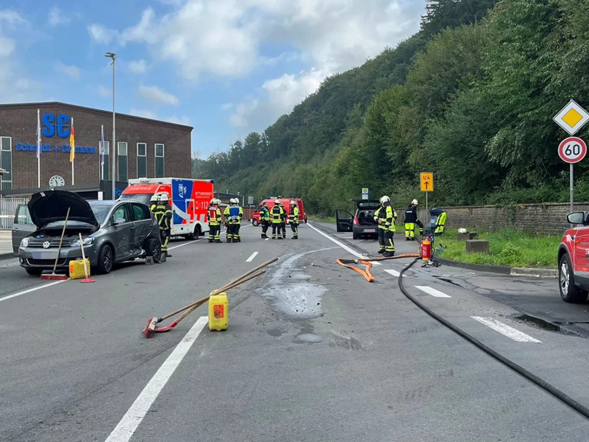 Auf einer Straße steht ein beschädigtes Fahrzeug mit offenen Türen, umgeben von Feuerwehrleuten. Ein Rettungswagen ist ebenfalls vor Ort.