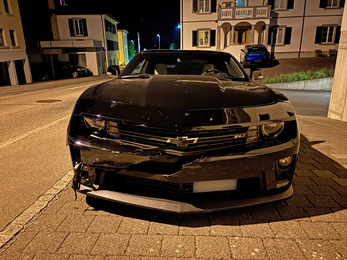 Ein schwarzes Auto mit Frontschäden steht nachts auf einer gepflasterten Straße. Im Hintergrund sind Wohngebäude zu sehen.