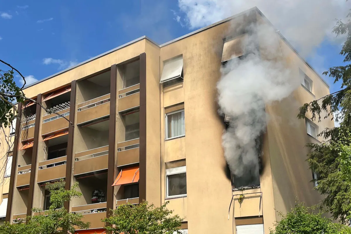 Ein beige- bis orangefarbenes Wohngebäude mit mehreren Etagen zeigt Rauch, der aus einem Fenster hervordringt. Der Rauch deutet auf ein mögliches Feuer in der Wohnung hin, während der Himmel im Hintergrund blau und am Tag klar ist.