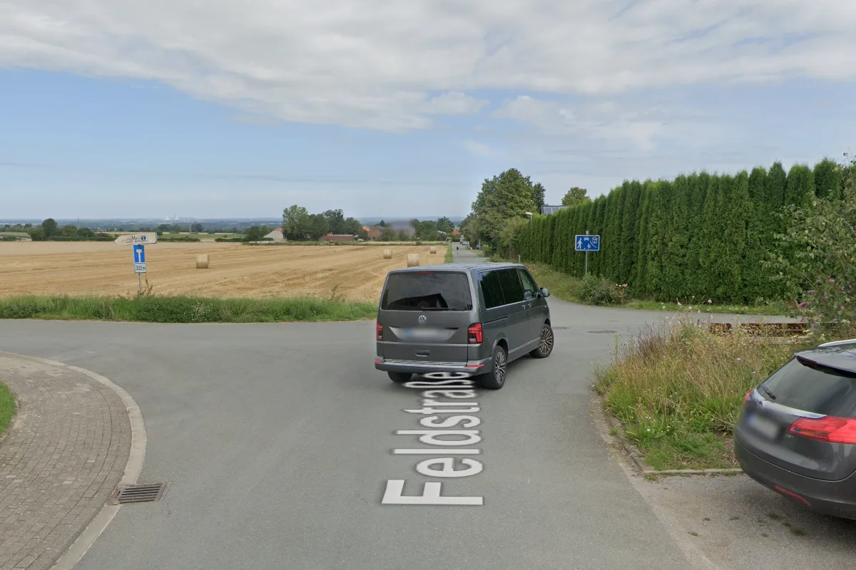 Ein grauer VW-Transporter fährt an einer Kreuzung auf einer Landstraße mit umliegenden Feldern und Bäumen.