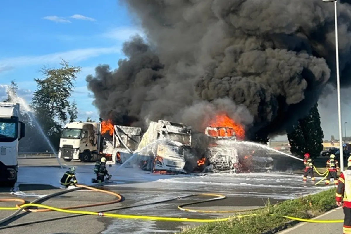 Mehrere LKWs stehen in Flammen, während Feuerwehrkräfte mit Schläuchen darauf löschen. Dichter schwarzer Rauch steigt auf.