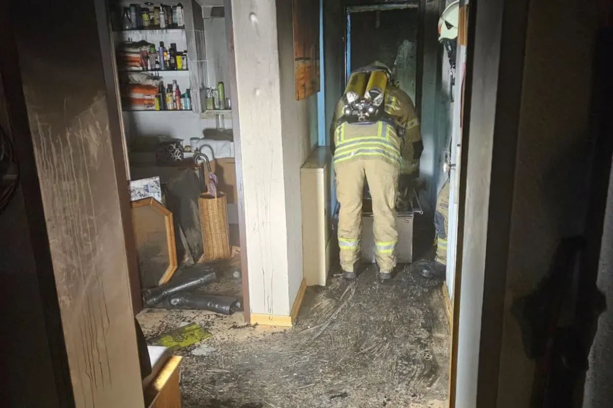 Ein Feuerwehrmann in voller Montur steht in einem verrauchten Raum. Der Boden ist mit Löschschaum bedeckt, Regale im Hintergrund sind voller Haushaltsgegenstände.