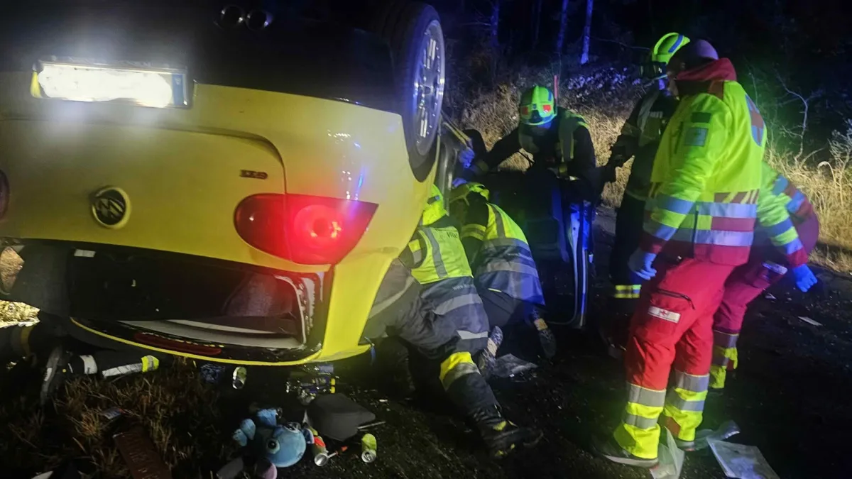 Mehrere Rettungskräfte sind nachts um ein umgekipptes gelbes Auto versammelt, um Insassen zu helfen. Sie tragen leuchtende Schutzkleidung.