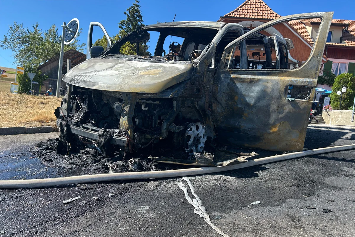 Ein komplett ausgebranntes Fahrzeug steht auf einer Straße, umgeben von verkohlten Überresten. Im Hintergrund sind ein Gebäude und blauer Himmel zu sehen.