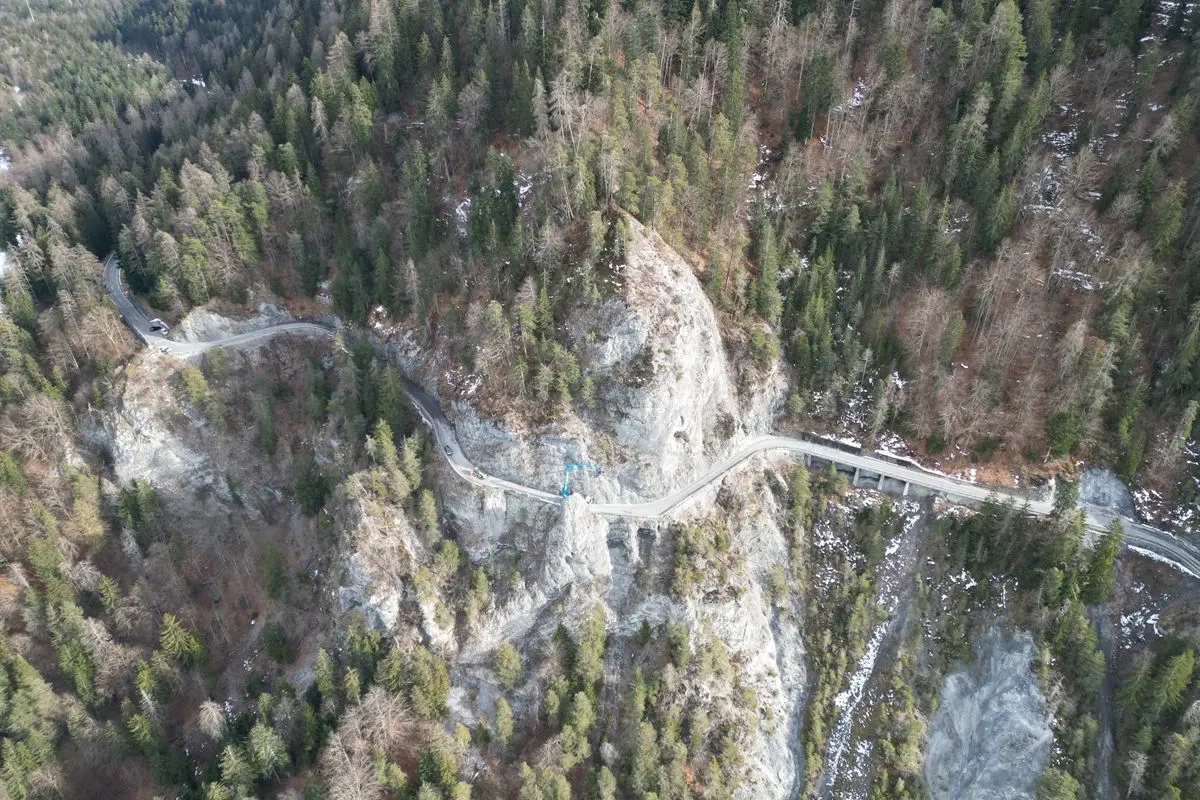 Die Luftaufnahme zeigt eine kurvenreiche Straße, die sich durch ein bergiges Waldgebiet schlängelt, mit kahlen Bäumen und Felsen.