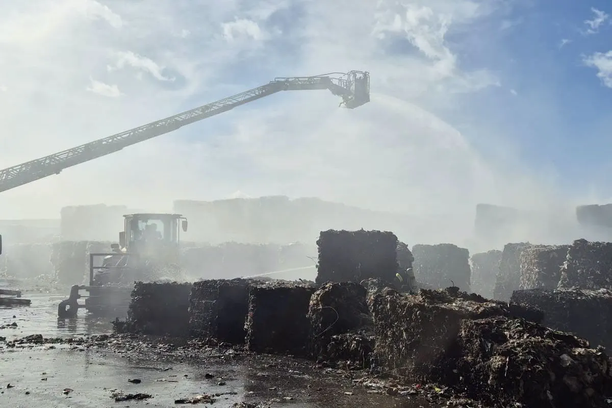 Ein Feuerwehrkran sprüht Wasser auf Müllballen, während Rauch über das Gelände zieht. Der Himmel ist blau mit einigen Wolken.