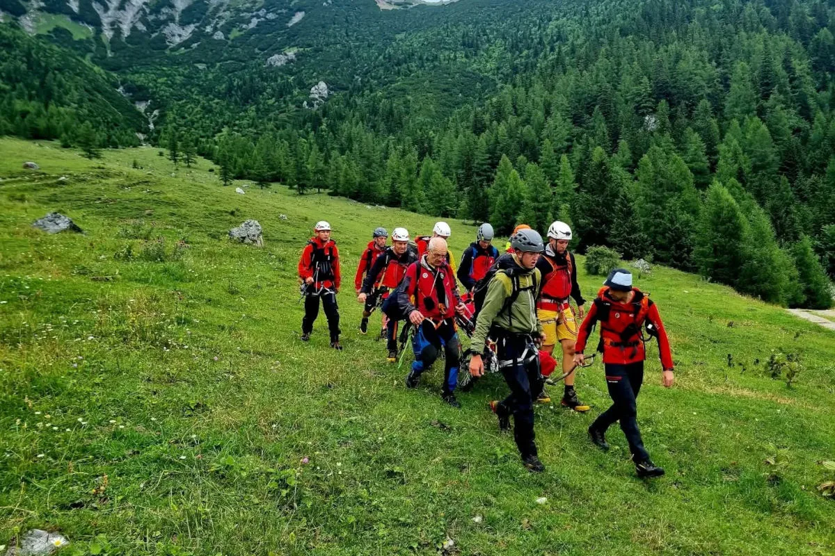 Ein Rettungsteam in Bergkleidung trägt eine Person auf einer Trage durch eine grüne, bergige Landschaft.