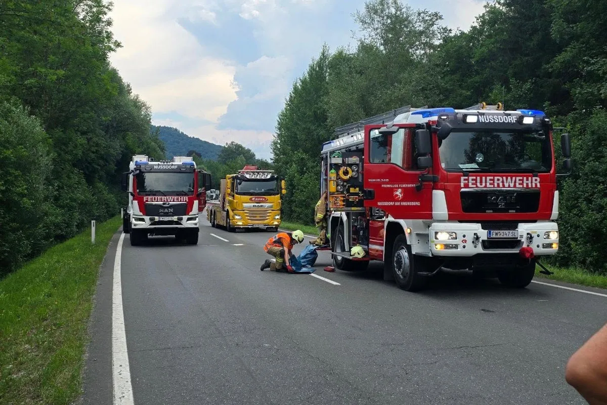 Feuerwehrfahrzeuge stehen auf einer Landstraße, ein Feuerwehrmann leistet einer Person auf der Straße Erste Hilfe.