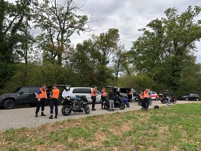 Eine Gruppe von Personen in leuchtenden Westen steht neben mehreren Fahrzeugen auf einer Straße, umgeben von Bäumen.