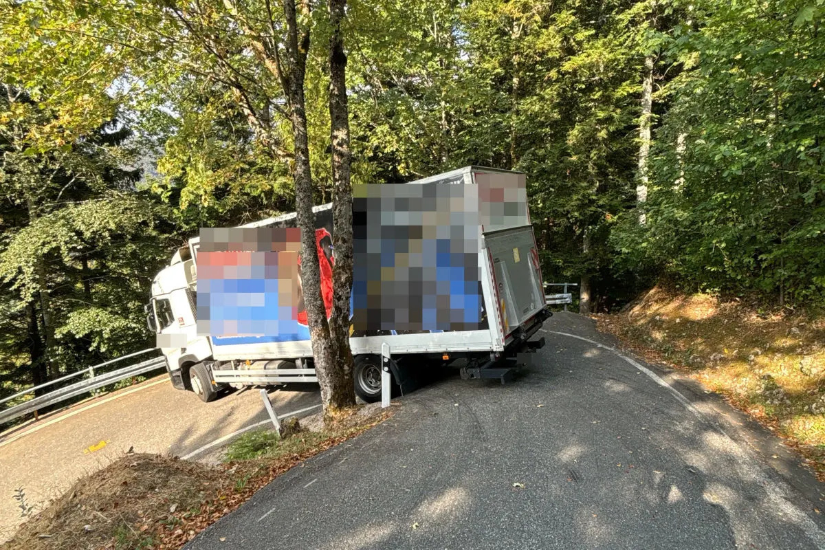 Ein weißer Lkw liegt quer auf einer kurvigen Straße, die durch ein Waldgebiet führt. Der Lkw blockiert die Straße komplett, umgeben von herbstlichem Laub.