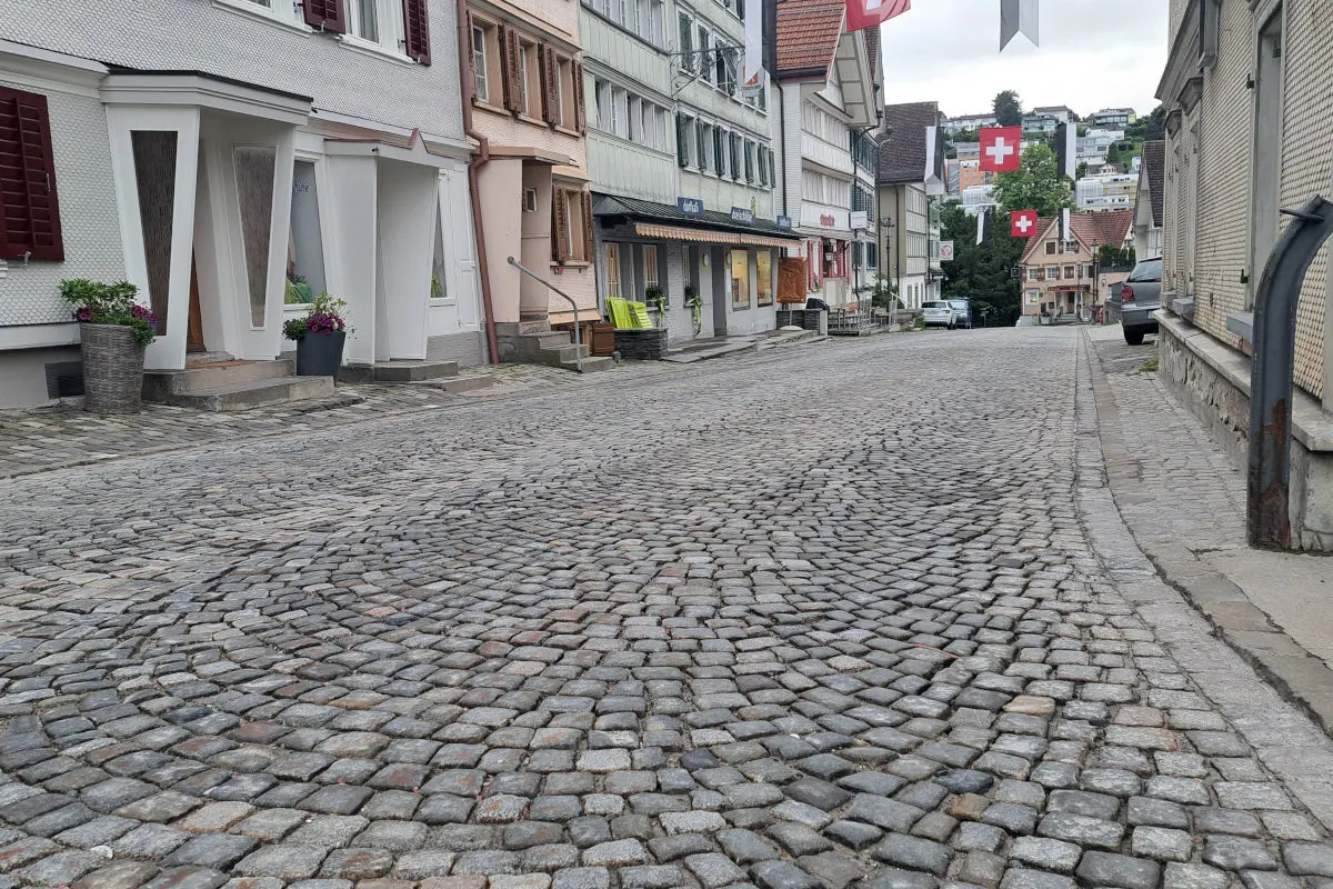 Eine leere Kopfsteinpflasterstraße führt durch eine Altstadt, gesäumt von Häusern mit Schweizer Fahnen. Die Szenerie wirkt ruhig und unbelebt.
