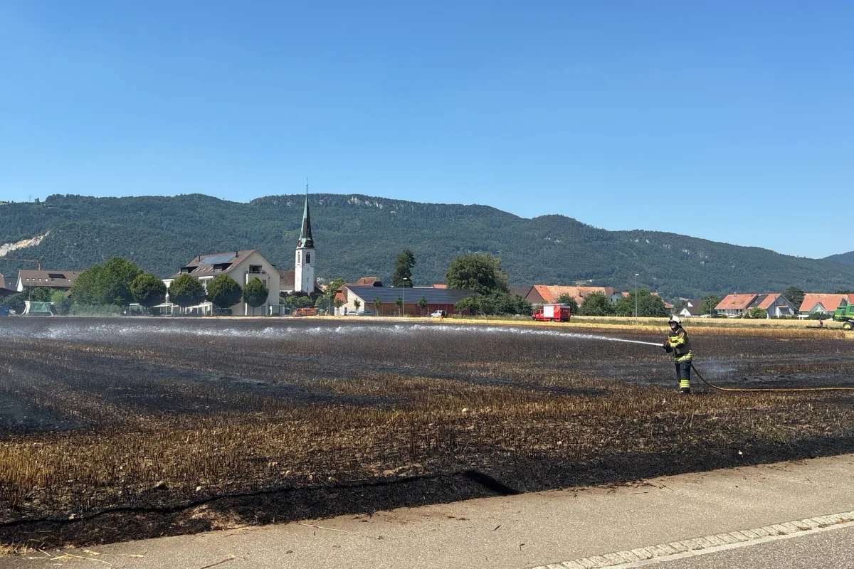 Ein Feuerwehrmann steht auf einem schwarzen, verbrannten Feld und löscht es mit einem Wasserschlauch. Im Hintergrund sind eine Kirche und einige Häuser sichtbar.