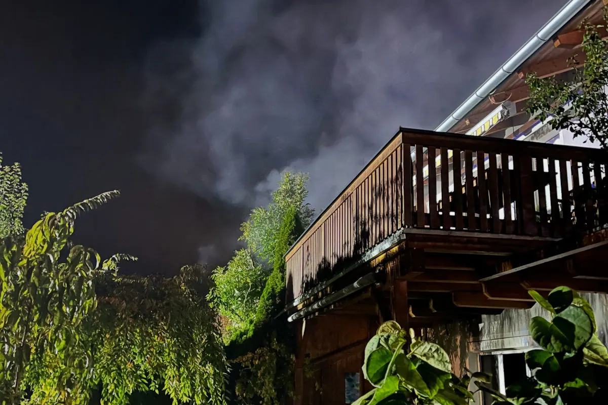 Ein Wohnhausbalkon bei Nacht, umgeben von Bäumen, zeigt aufsteigenden Rauch in der Dunkelheit. Der Himmel ist dunkel und wolkig.
