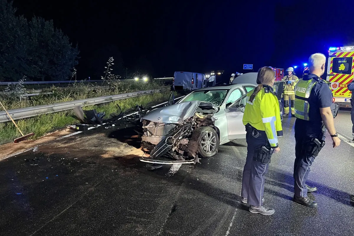 Ein Auto mit stark beschädigter Front steht auf einer Autobahn. Im Hintergrund sind Polizisten und Rettungskräfte sichtbar. Die Szene ist nachts beleuchtet.