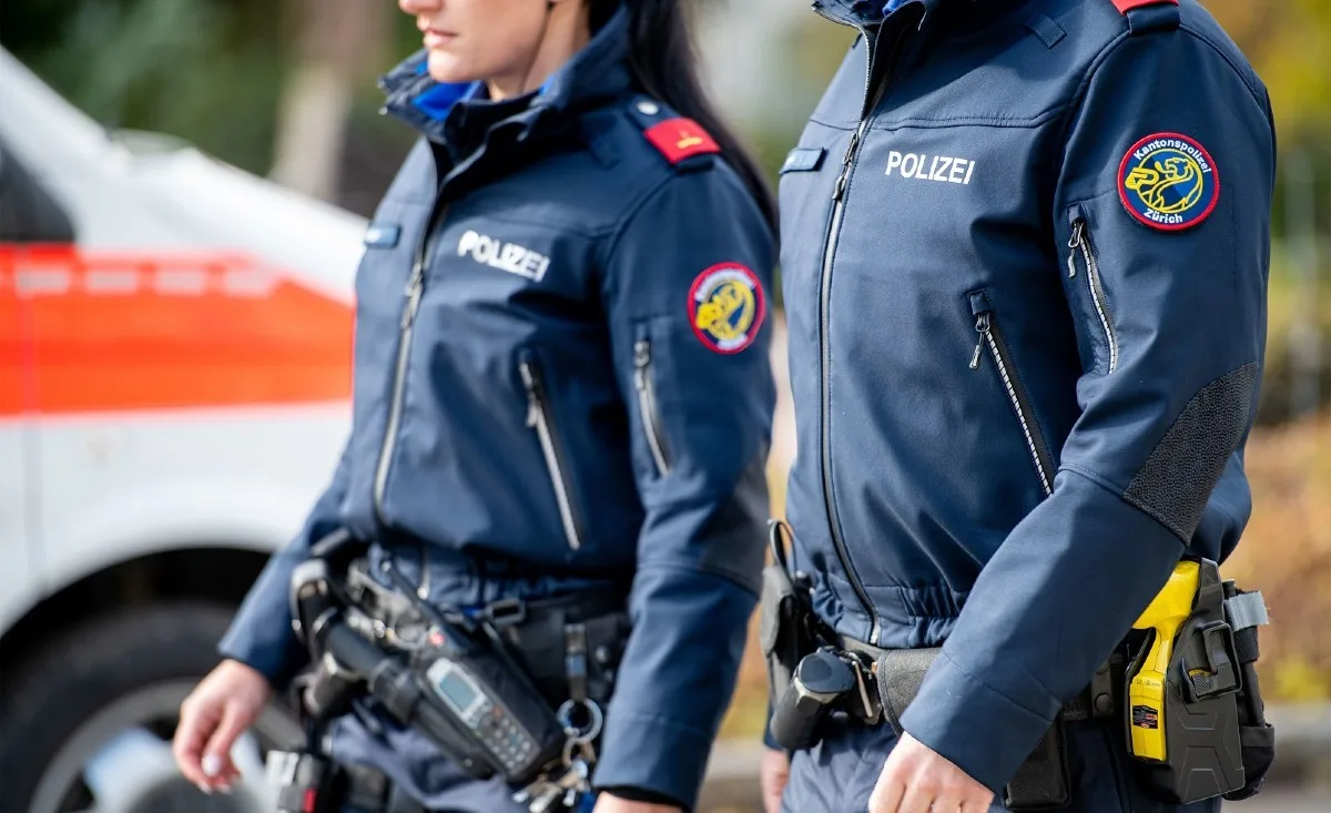 Zwei Polizisten in blauen Uniformen, erkennbar an den Aufschriften 'Polizei' und dem Wappen der Kantonspolizei Zürich. Im Hintergrund ist ein Polizeiwagen sichtbar.