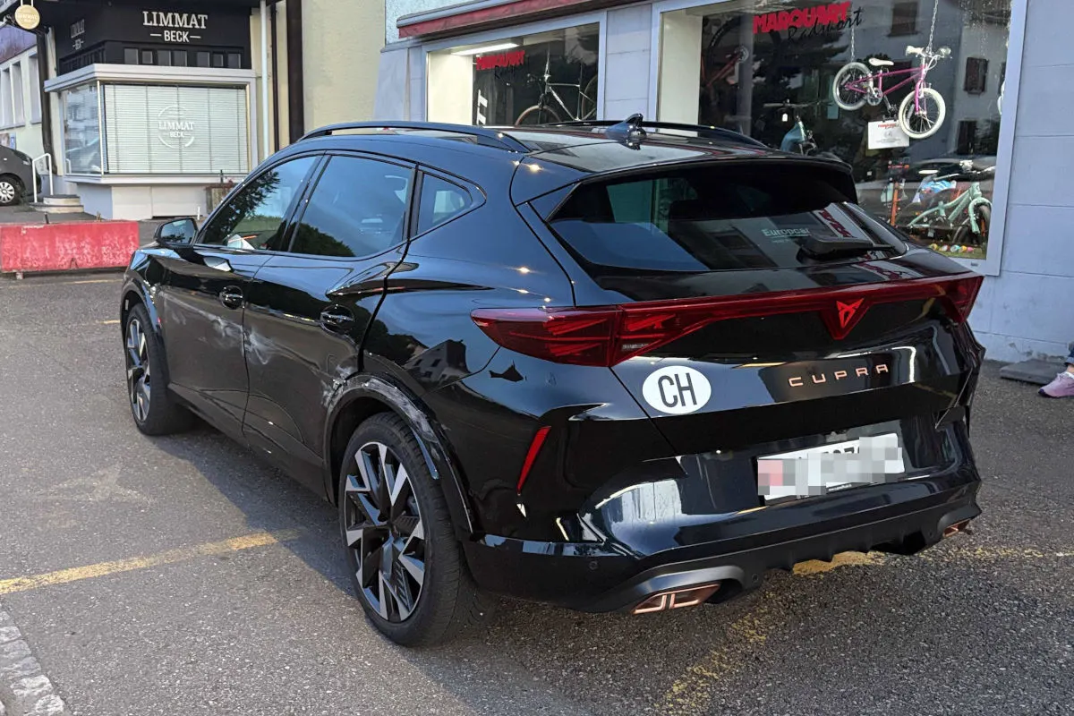 Ein schwarzes Auto steht auf einem Parkplatz vor einem Geschäft. Der Wagen hat einen CH-Aufkleber am Heck.
