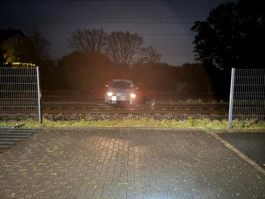 Ein Auto ist zwischen Bahngleisen eingeklemmt. Es ist dunkel, und das Fahrzeug wird von beiden Seiten durch Zäune begrenzt.