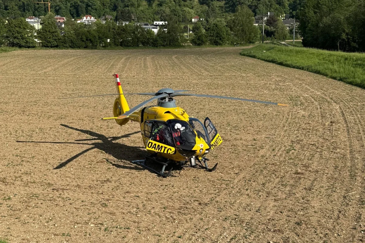 Ein gelber Rettungshubschrauber mit der Aufschrift 'ÖAMTC' steht auf einem braunen Feld. Im Hintergrund sind Bäume und Wohnhäuser zu sehen.