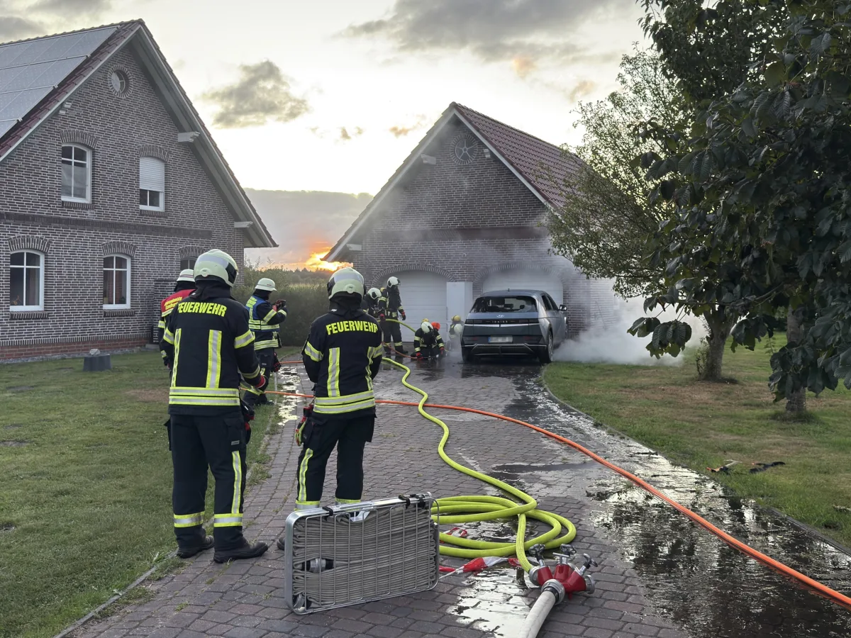 Mehrere Feuerwehrleute sind vor einem brennenden Auto in einer Garage zu sehen, während sie das Feuer löschen. Im Vordergrund liegen Schläuche und Ausrüstung.