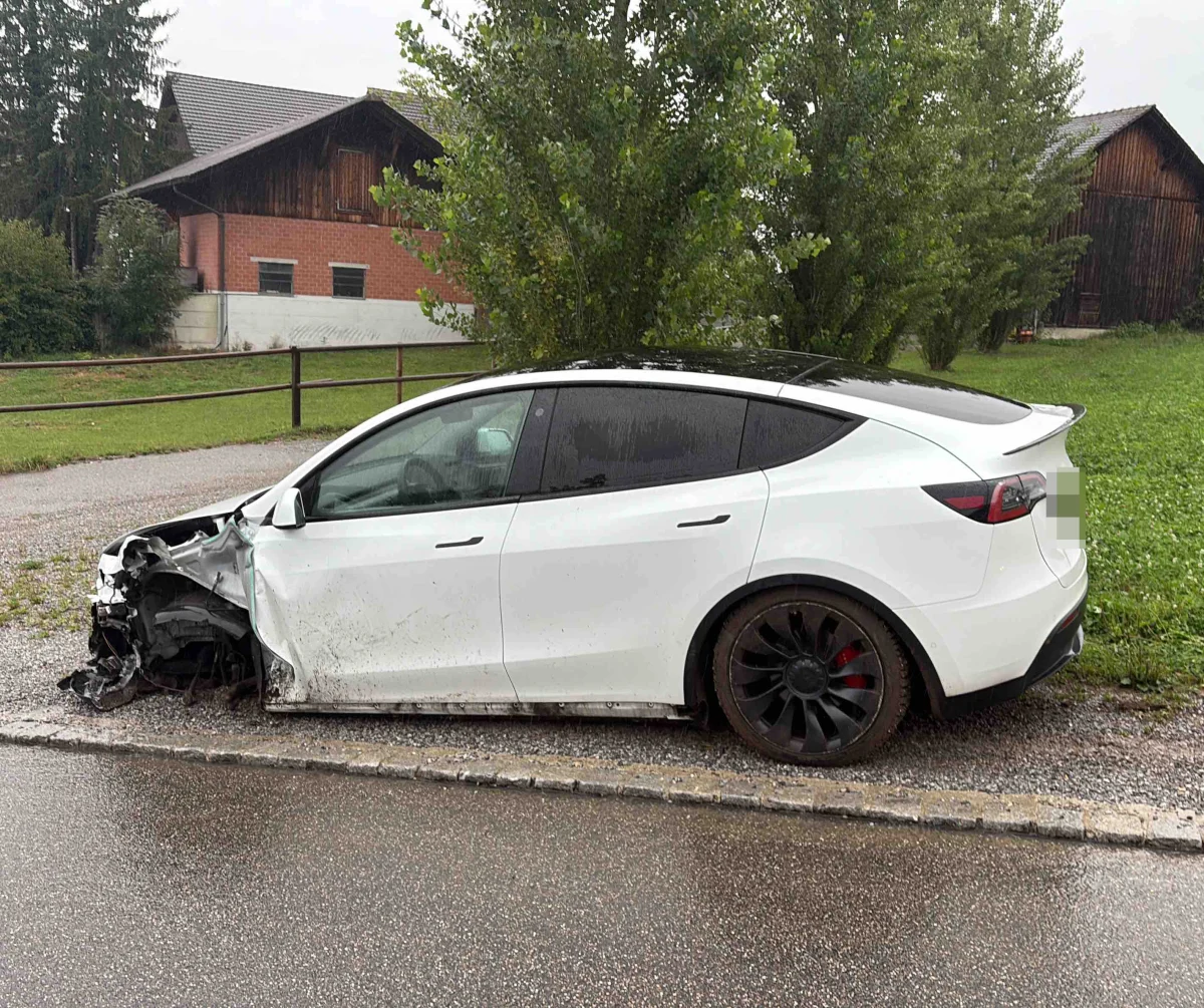 Ein weißes Auto steht mit einem erheblichen Schaden an der Front am Straßenrand. Im Hintergrund sind ein Haus und Bäume zu sehen.