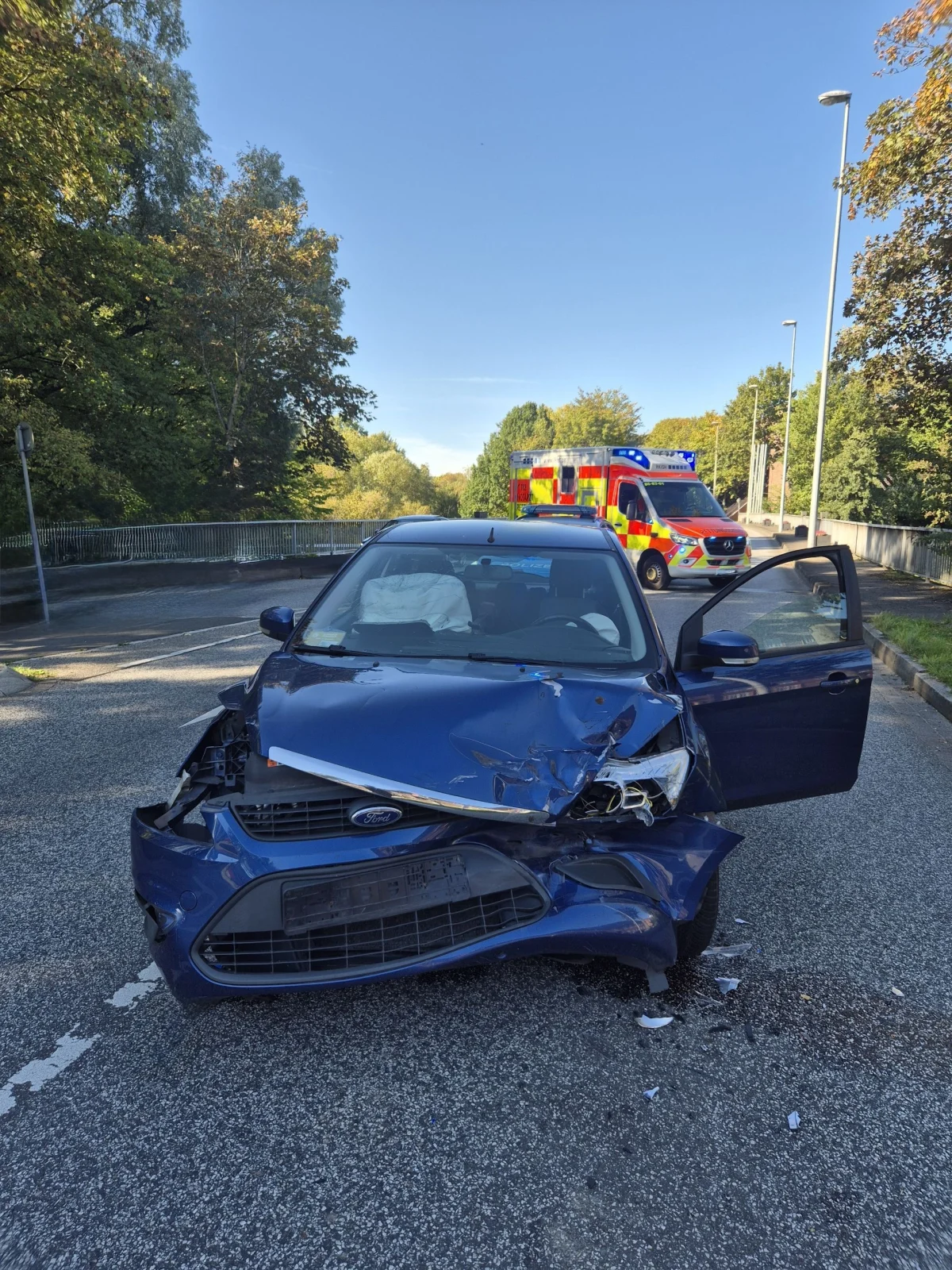 Ein stark beschädigtes blaues Fahrzeug steht auf einer Straße. Im Hintergrund ist ein Rettungswagen zu sehen.