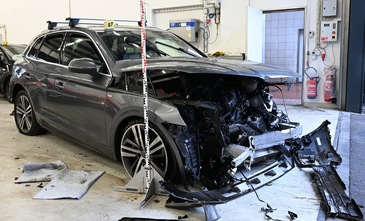 Das Bild zeigt ein stark beschädigtes graues Auto, das in einer Werkstatt steht. Die Front des Fahrzeugs ist erheblich beschädigt.