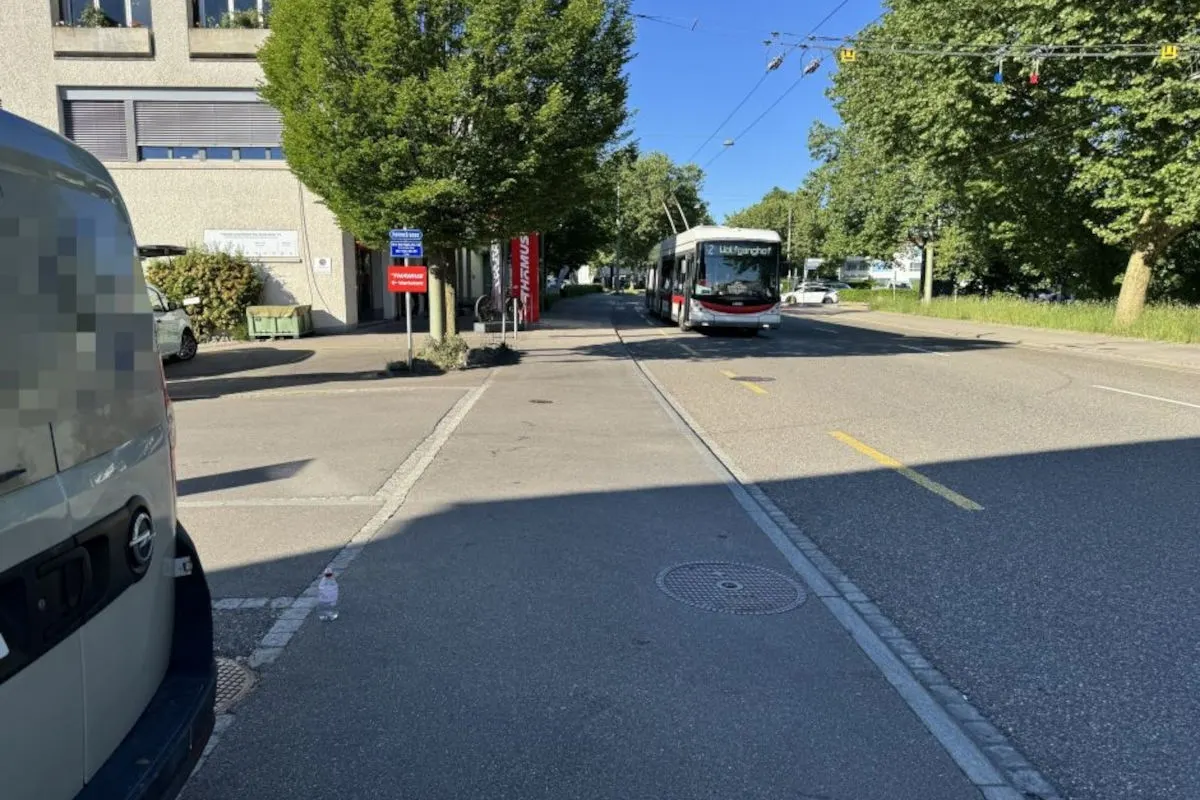 Ein Bus bewegt sich auf einer Stadtstraße, umgeben von Bäumen und einem Gebäude. Die Szene ist hell und sonnig.