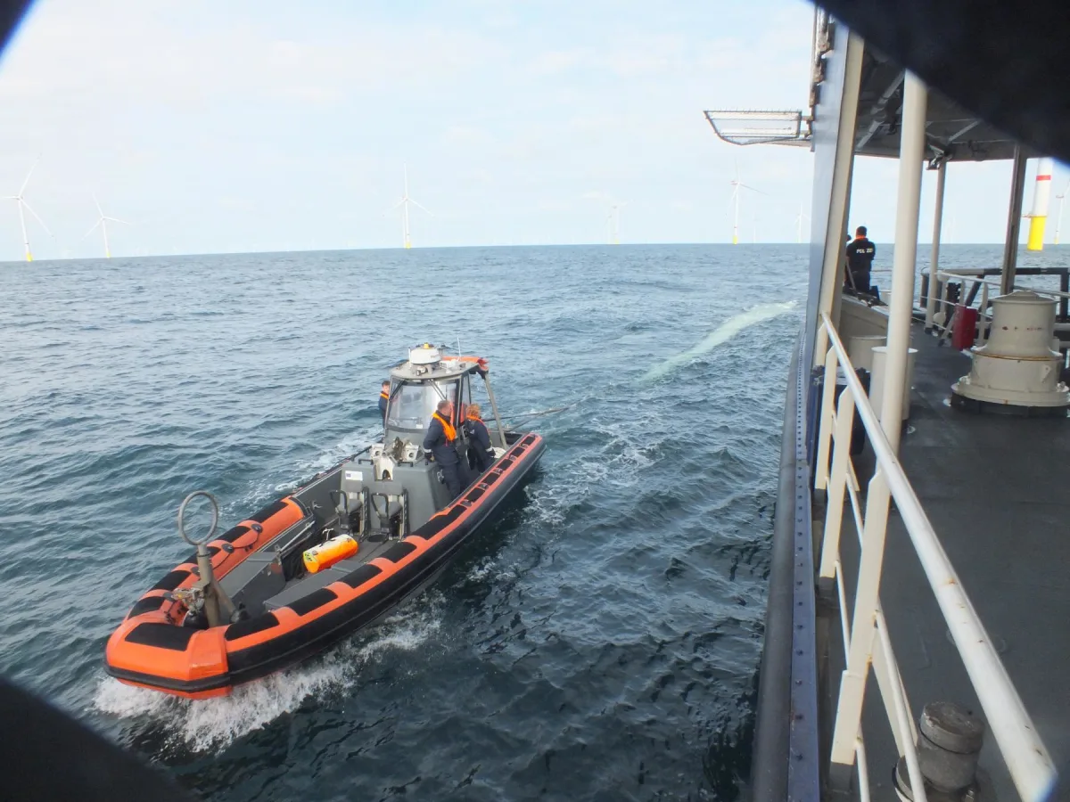 Ein kleines Patrouillenboot fährt neben einem größeren Schiff auf der offenen See, im Hintergrund sind Windkraftanlagen sichtbar.